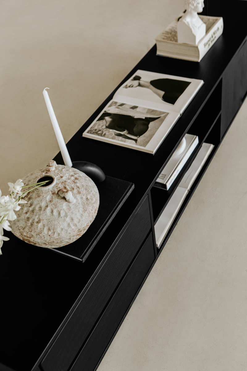 The Indre Media Console displays a textured vase with white flowers, a white candle in a holder, an open art book, and stacked books on a light floor for a refined minimalist look.