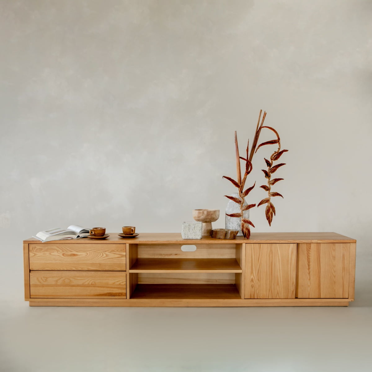 The Indre Media Console features drawers and shelves for ample storage, styled with dried plants in a vase, stacked wooden bowls, cups, and an open book, all against a neutral backdrop for a modern touch.