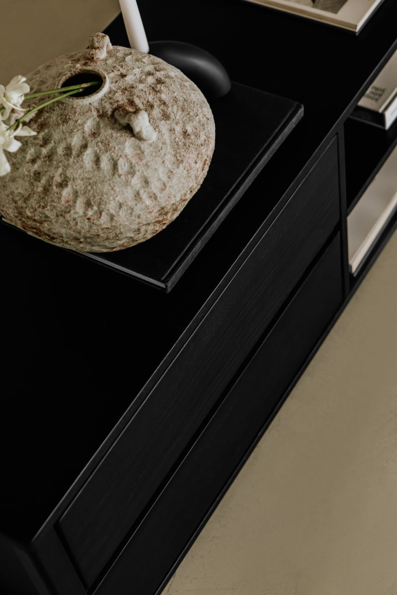 The Indre Media Console, featuring shelves for storage, displays a large stone vase with lid and white flowers, plus a candle on top—all set on a beige floor.