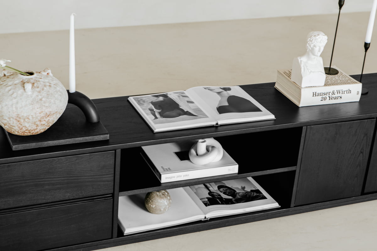 The Indre Media Console in dark wood showcases candles, art books (some open to black-and-white images), a small sculpture, and a Greco-Roman style bust, all accented by minimalist design and neutral-toned decor.