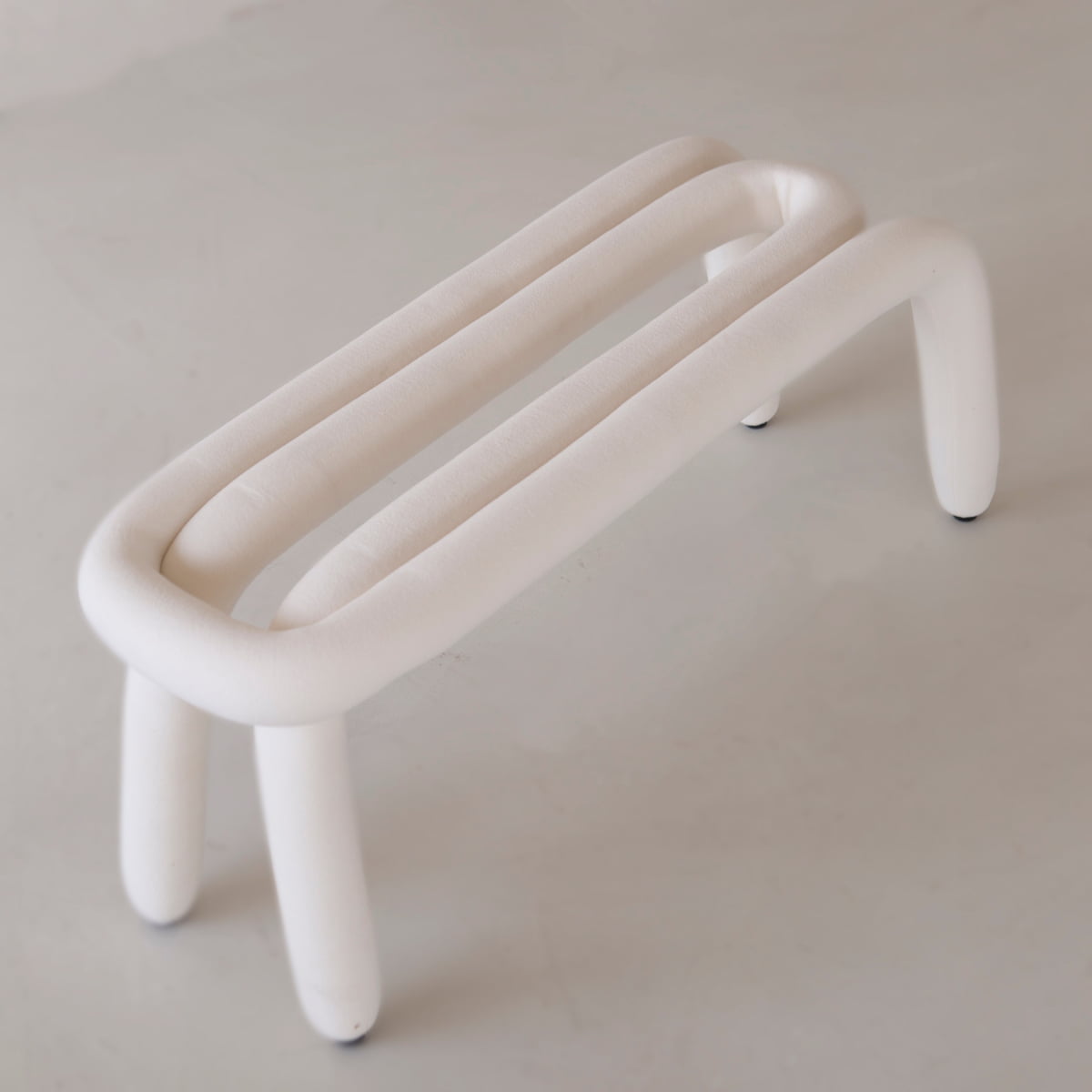 The Loud Bench features a minimalist, modern design with a soft white polyurethane foam-upholstered tubular frame, four rounded legs, and two parallel cushion tubes for an open, airy look on a light-colored floor.