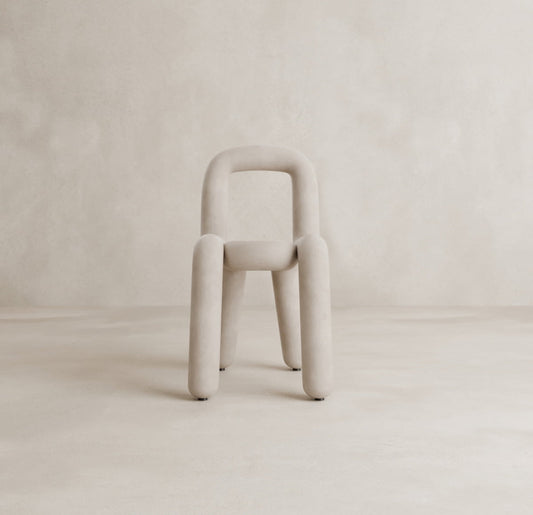 The Loud Chair is a minimalist beige seat with a rounded, sculptural design and thick tubular legs and backrest crafted from polyurethane foam, shown against a plain, light-colored textured background.