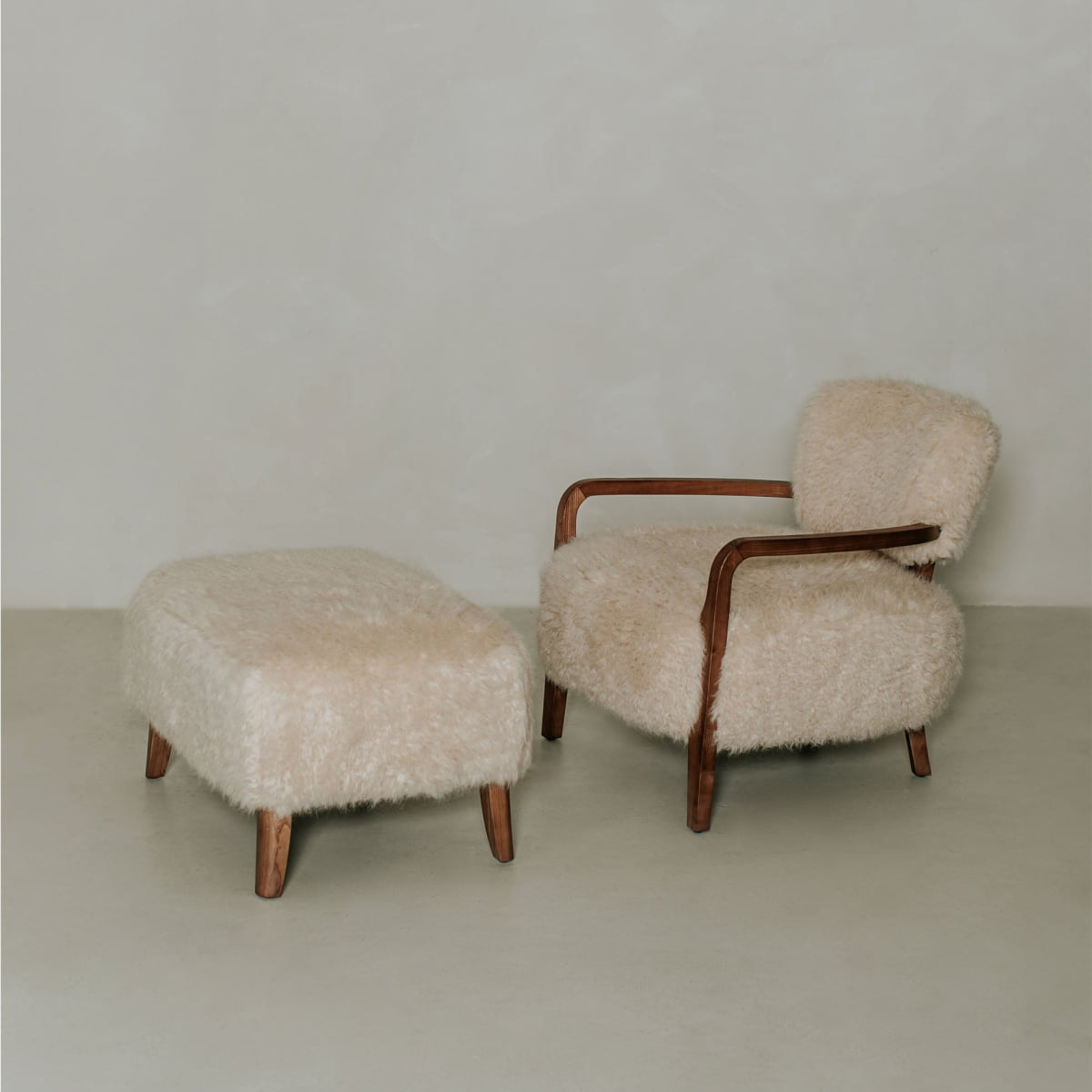 The Malmo Chair with Ottoman features wooden armrests and legs and is upholstered in cream shearling, creating minimalist sophistication against a light wall and floor.