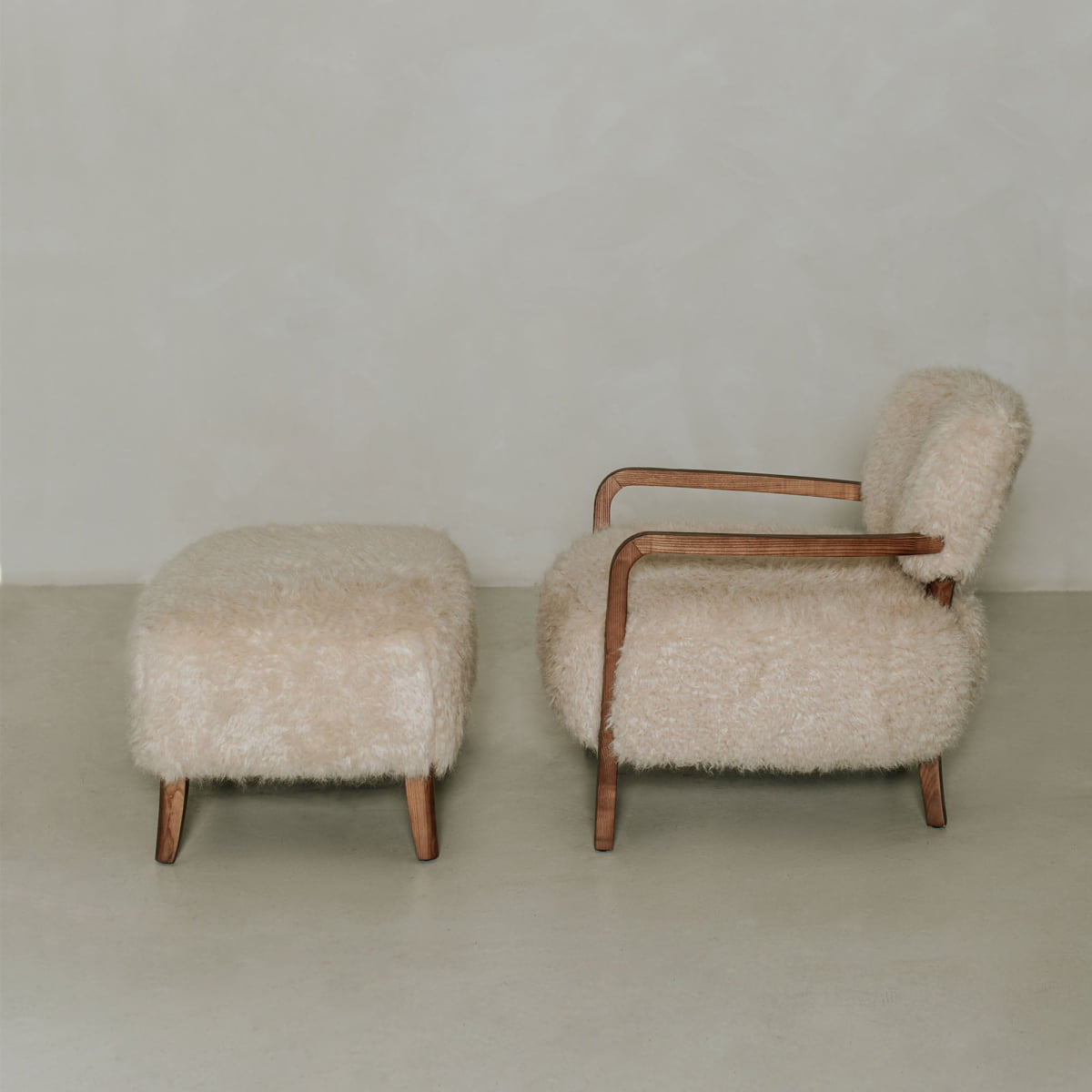 The Malmo Chair with Ottoman features plush cream shearling upholstery and wooden armrests, exemplifying minimalist sophistication against a light background.