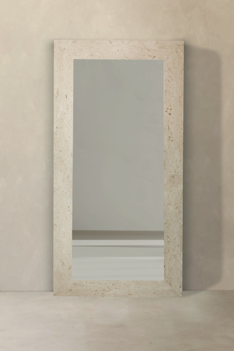 The Marbella Floor Mirror is a full-length rectangular mirror with a travertine stone frame, shown leaning against a textured light wall and reflecting a bare wall with a white baseboard on the smooth floor.