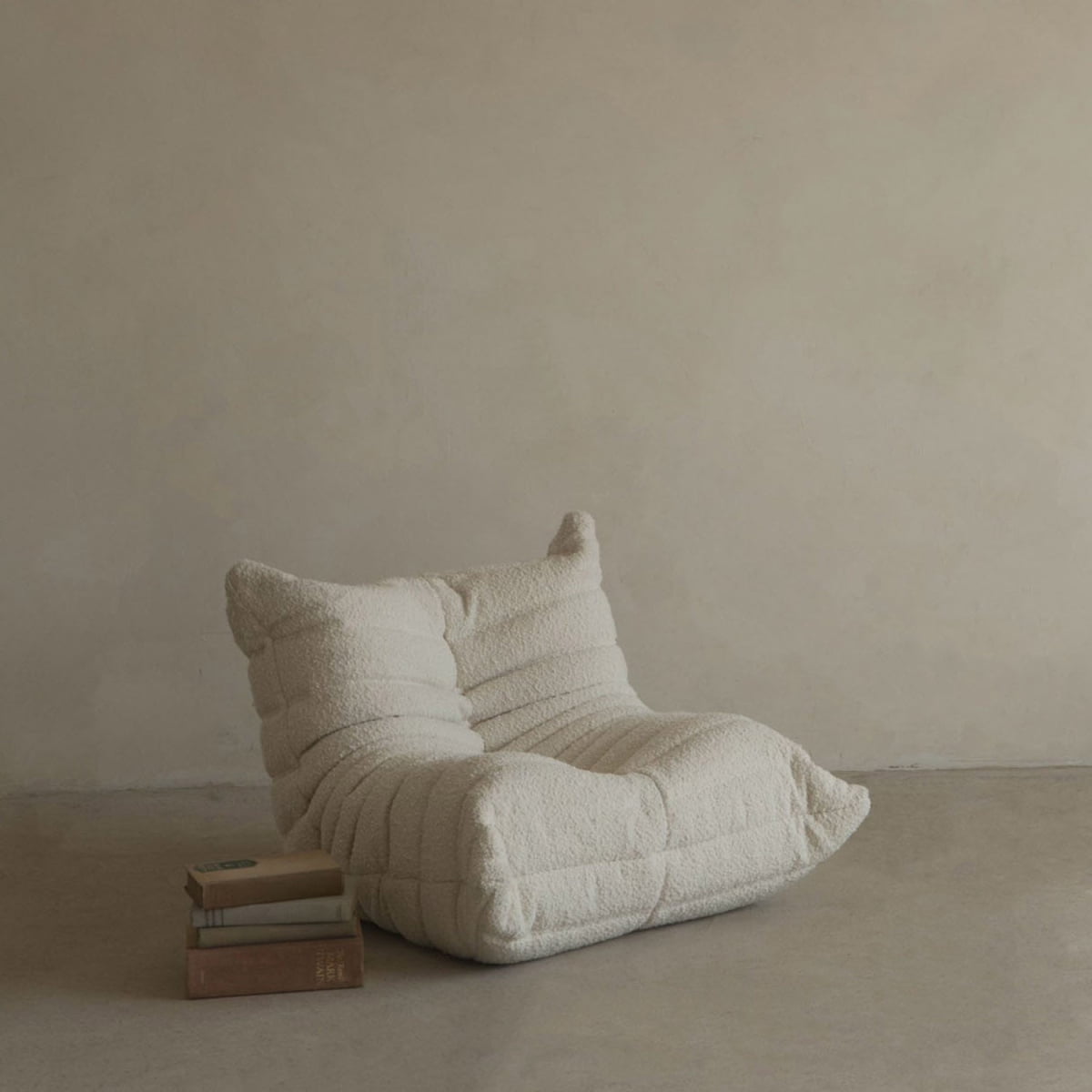 The cream Mini Rolo Lounge Chair, with its cozy textured design, sits on a concrete floor beside books and a beige wall—an ideal choice for a minimalist playroom or child’s space.