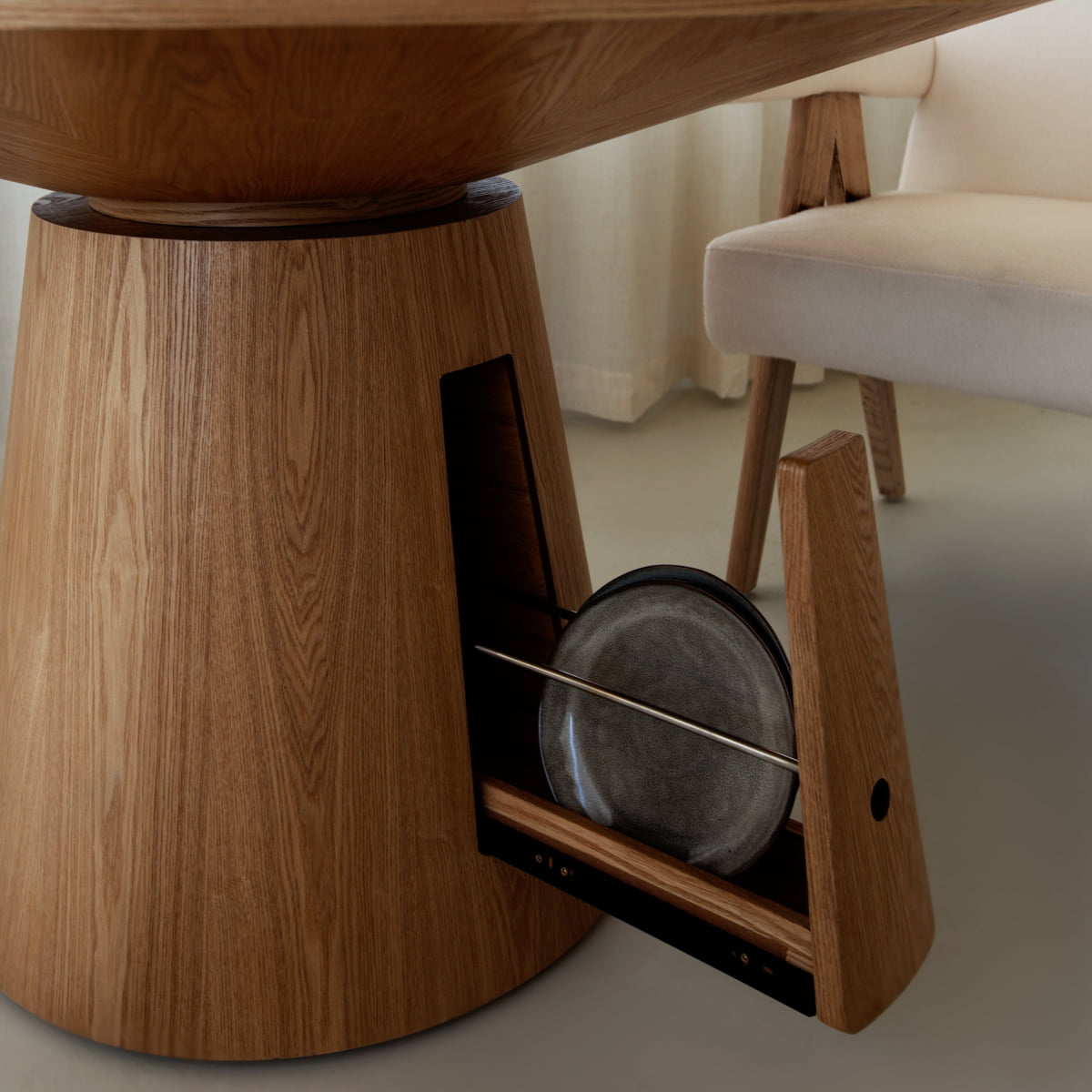 A close-up of the Nola + Elsa Dining Set shows its round wood table base with a hidden storage slot holding two gray plates, beside a beige upholstered chair on a light-colored floor.