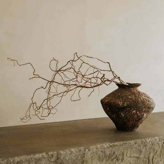 The Oran, a textured dark brown ceramic vase from our artisan collection, holds twisted branches on a rough stone surface against a beige wall—perfect for rustic vases and timeless home decor.