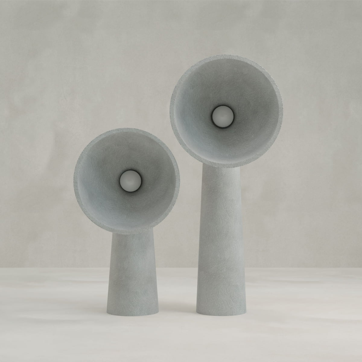 The Orbis Medium Floor Lamps, featuring minimalist gray cone-shaped designs with wide, flared tops, stand on a neutral surface against a light gray backdrop, highlighting sculptural artistry and an organic stone-like aesthetic.