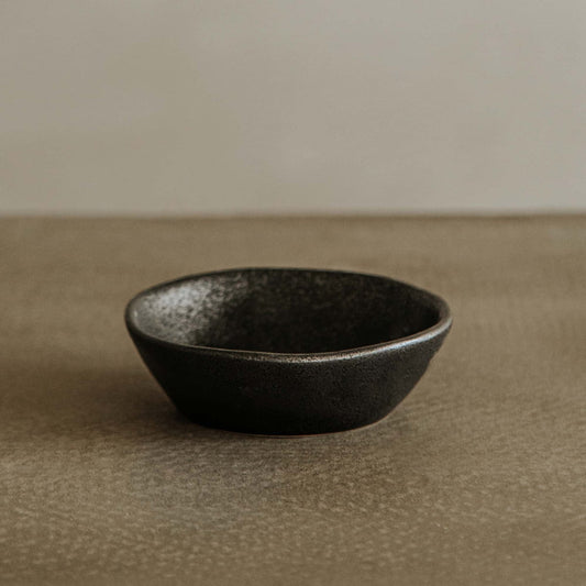 The Pebble Speckled Bowl Large, featuring a sleek black ceramic design, rests on a textured brown surface with a light beige background.