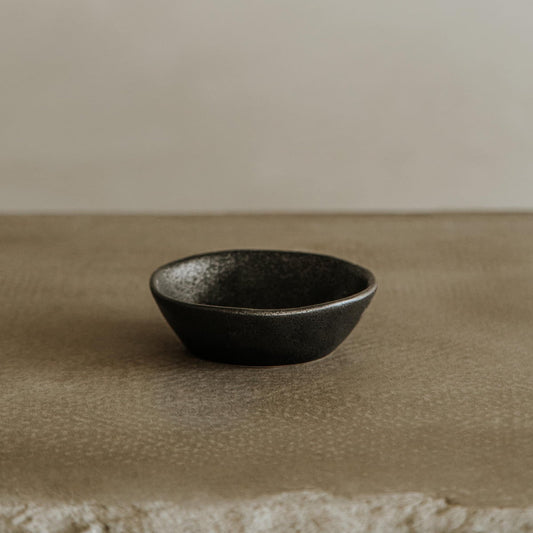The Pebble Speckled Bowl Medium, a small black ceramic bowl, rests on a textured neutral surface with a soft beige background.