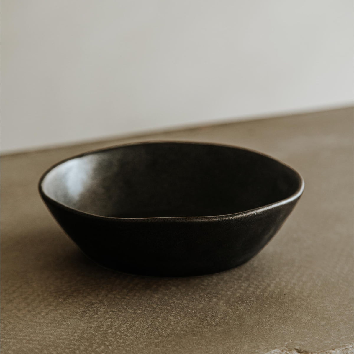 The Pebble Speckled Bowl Small, featuring a simple round design in dark ceramic, sits on a textured beige surface against a plain light background.