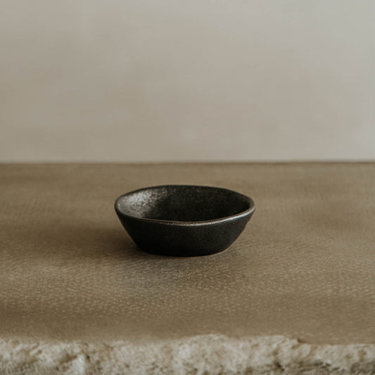 The Pebble Speckled Bowl Small, empty and dark, rests on a textured beige surface against a neutral background.