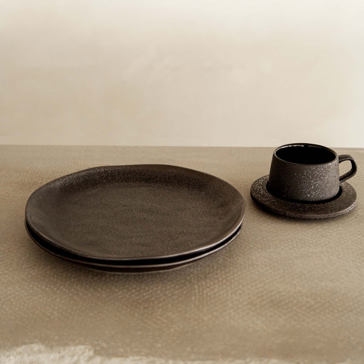 A Pebble Speckled Dish Large is stacked with another plate on the left, beside a matching dark ceramic cup and saucer to the right, all arranged on a neutral textured surface.