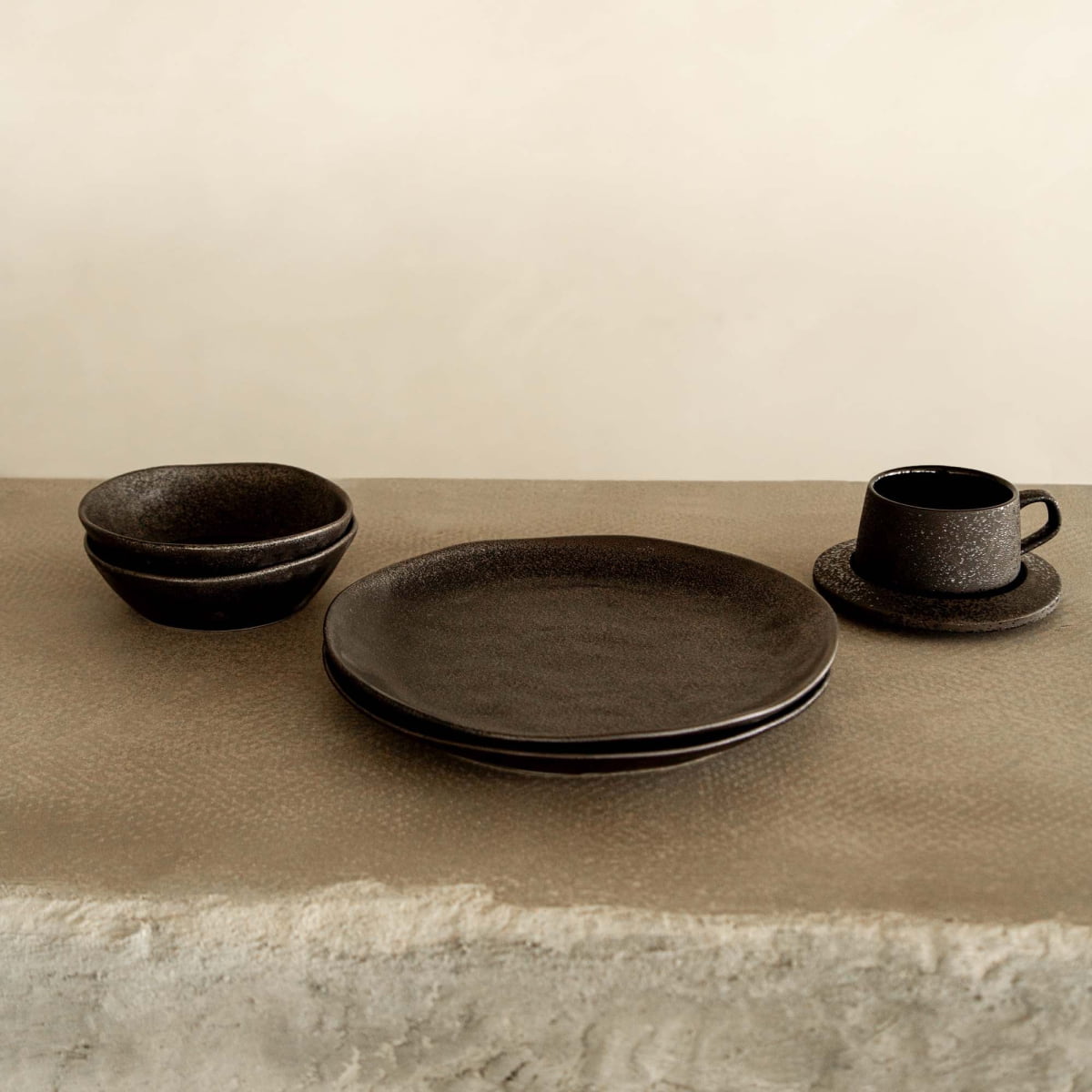 A Pebble Speckled Dish Large in black ceramic, paired with two stacked plates and a cup with saucer, is displayed on a rough, stone-like surface against a plain beige background.