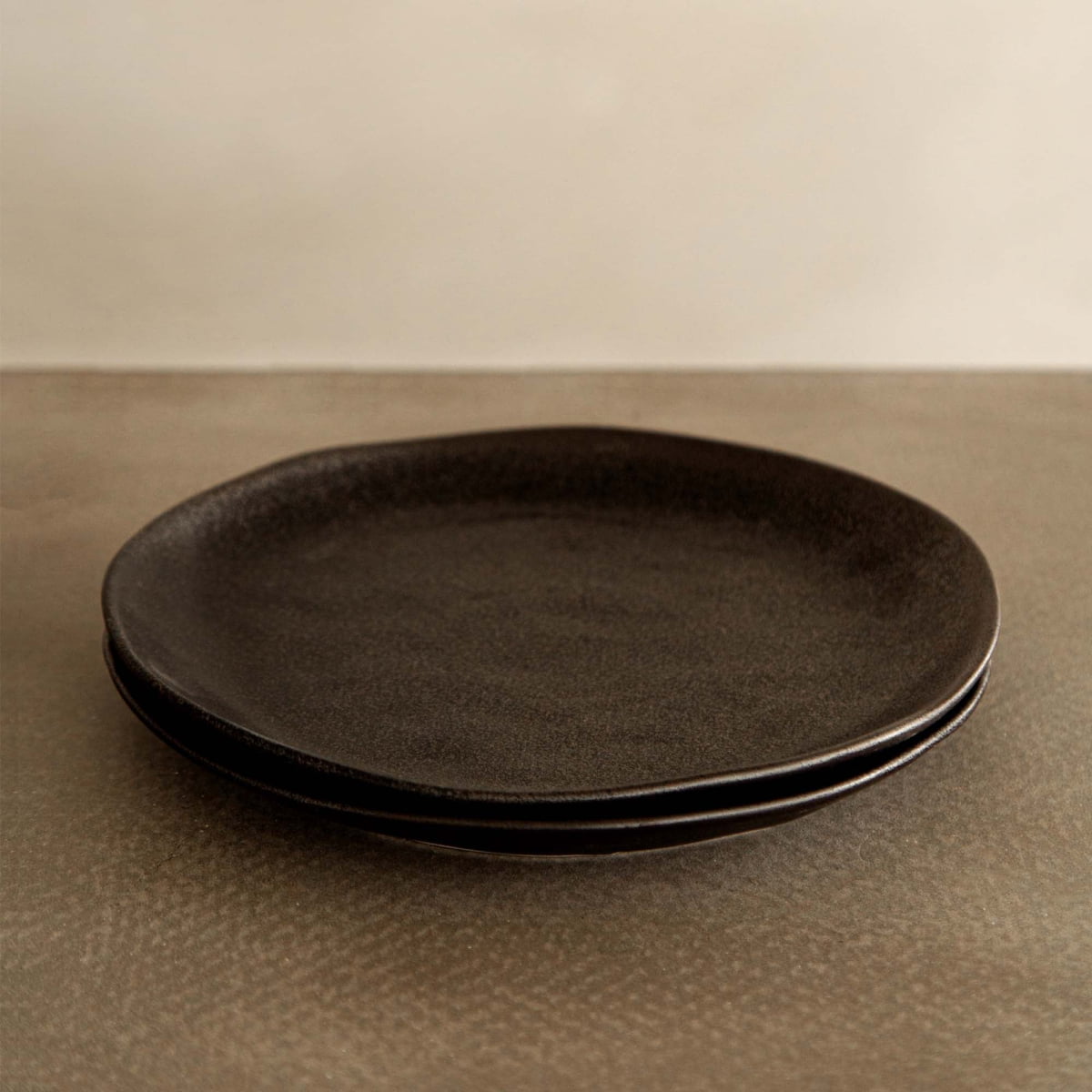 Two Pebble Speckled Dish Large plates, dark and round, are stacked together on a textured brown surface with a neutral beige background.
