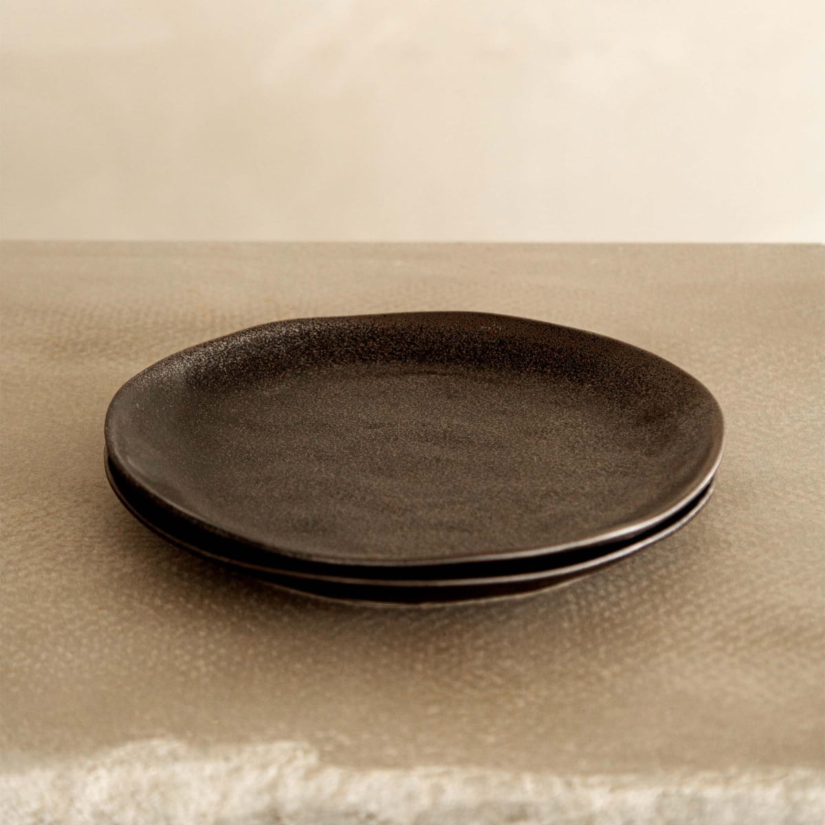 Two Pebble Speckled Dish Large plates are stacked, showcasing their dark, round ceramic design on a textured, light-colored surface with a neutral background.