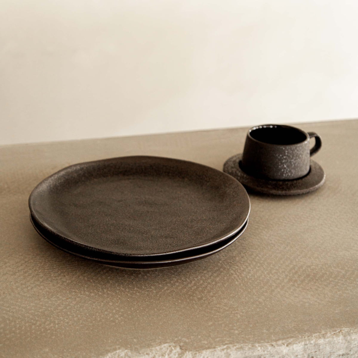 Two Pebble Speckled Dish Large plates are stacked on a textured surface next to a matching cup and saucer, all set against a minimalist, neutral-toned background.