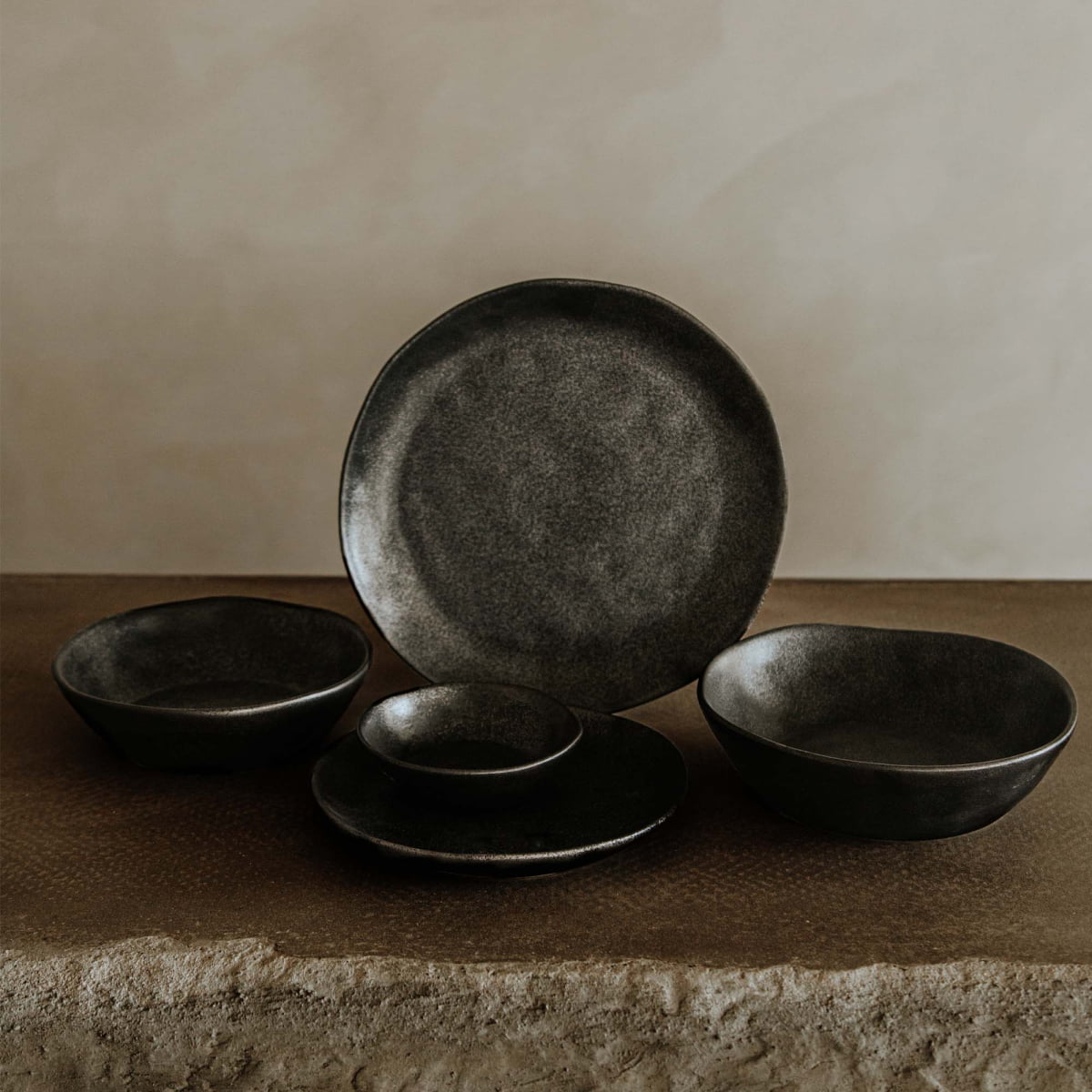 A Pebble Speckled Bowl Medium, part of a dark matte ceramic dinnerware set, is displayed on a textured stone surface with a neutral beige background.