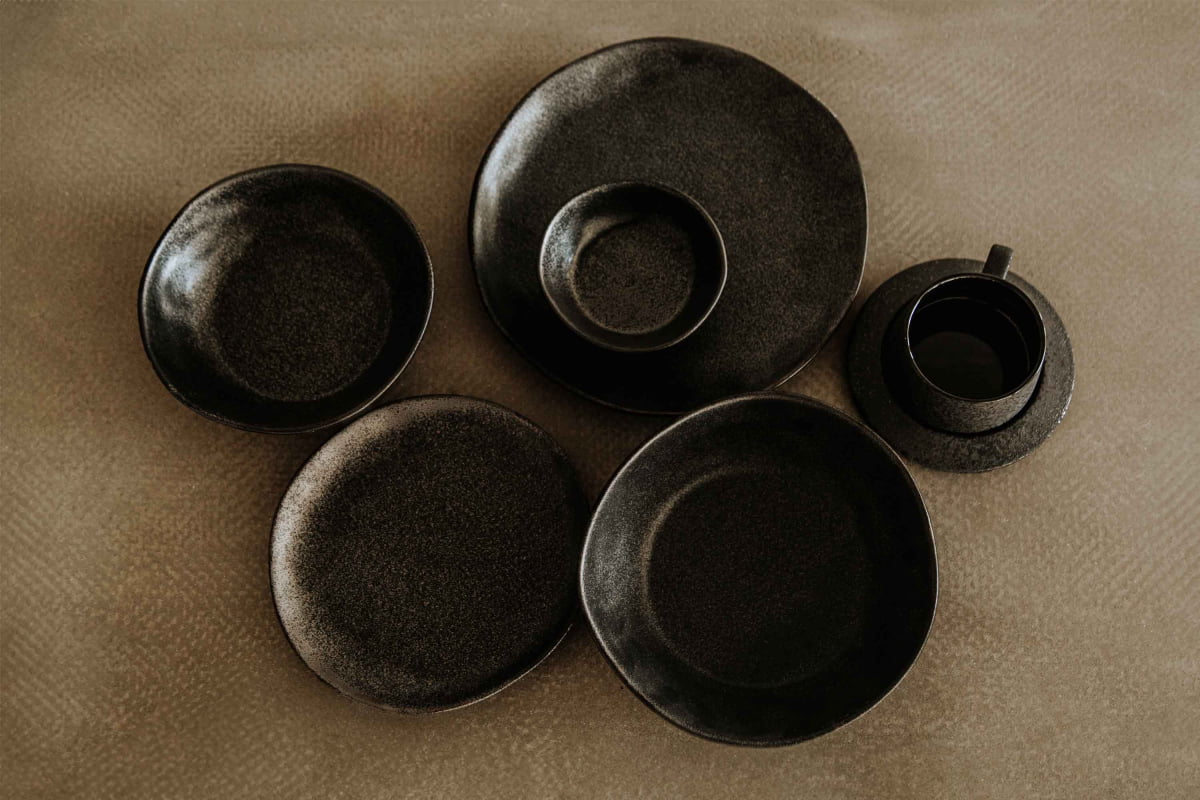 A Pebble Speckled Bowl Medium, part of a set with five dark ceramic dishes and a matching cup, all featuring an organic, handmade look, is arranged on a textured brown surface.