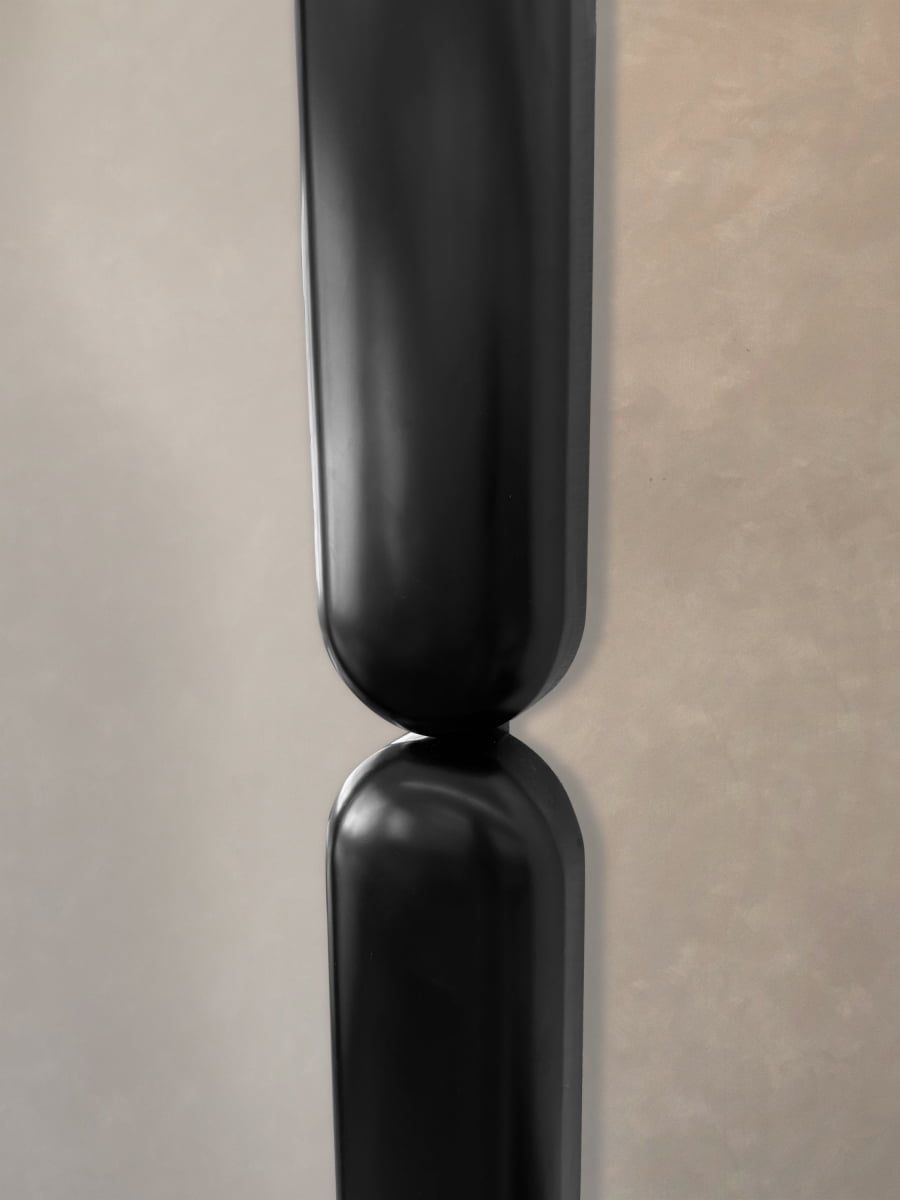Close-up of two Pitti Mirror Large In Black pieces, their smooth, reflective surfaces aligned end to end against a beige textured wall, creating a minimalist and modern look with soft shadows.