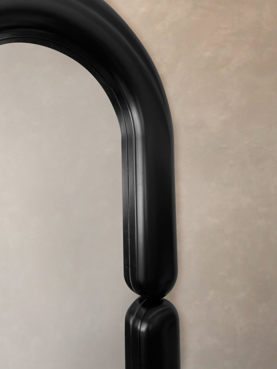 Close-up of the Pitti Mirror Large in Black, featuring a modern round reflective surface and black rounded arch, set against a textured beige wall to emphasize its smooth lines and minimalist design.
