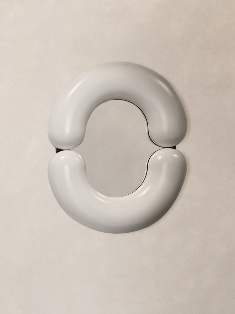 The Pitti Mirror Small In White is a wall-mounted oval mirror with two curved, padded white cushions at its top and bottom, creating an abstract, modern look against a plain beige background.