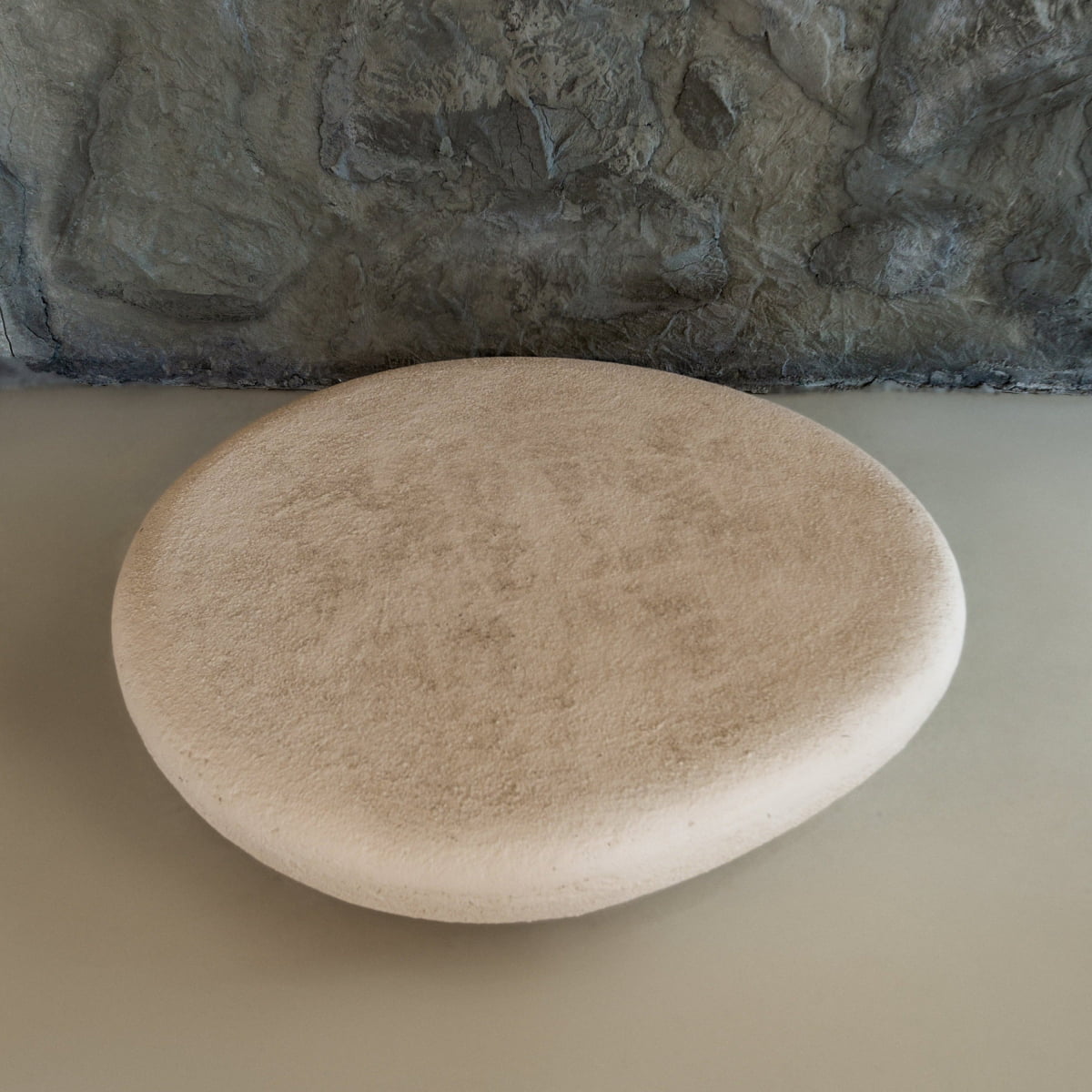 The Pluto Coffee Table, with its round beige stone-like form and smooth, minimalist design, sits on a light floor before a gray stone wall, serving as a chic modern centerpiece for any space.