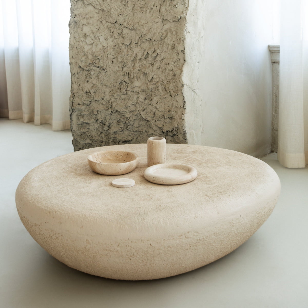 The Pluto Coffee Table, a round textured beige centerpiece, is styled with minimalist stone bowls and a small vase in a bright room with white curtains and an unfinished wall, bringing modern aesthetics to the space.