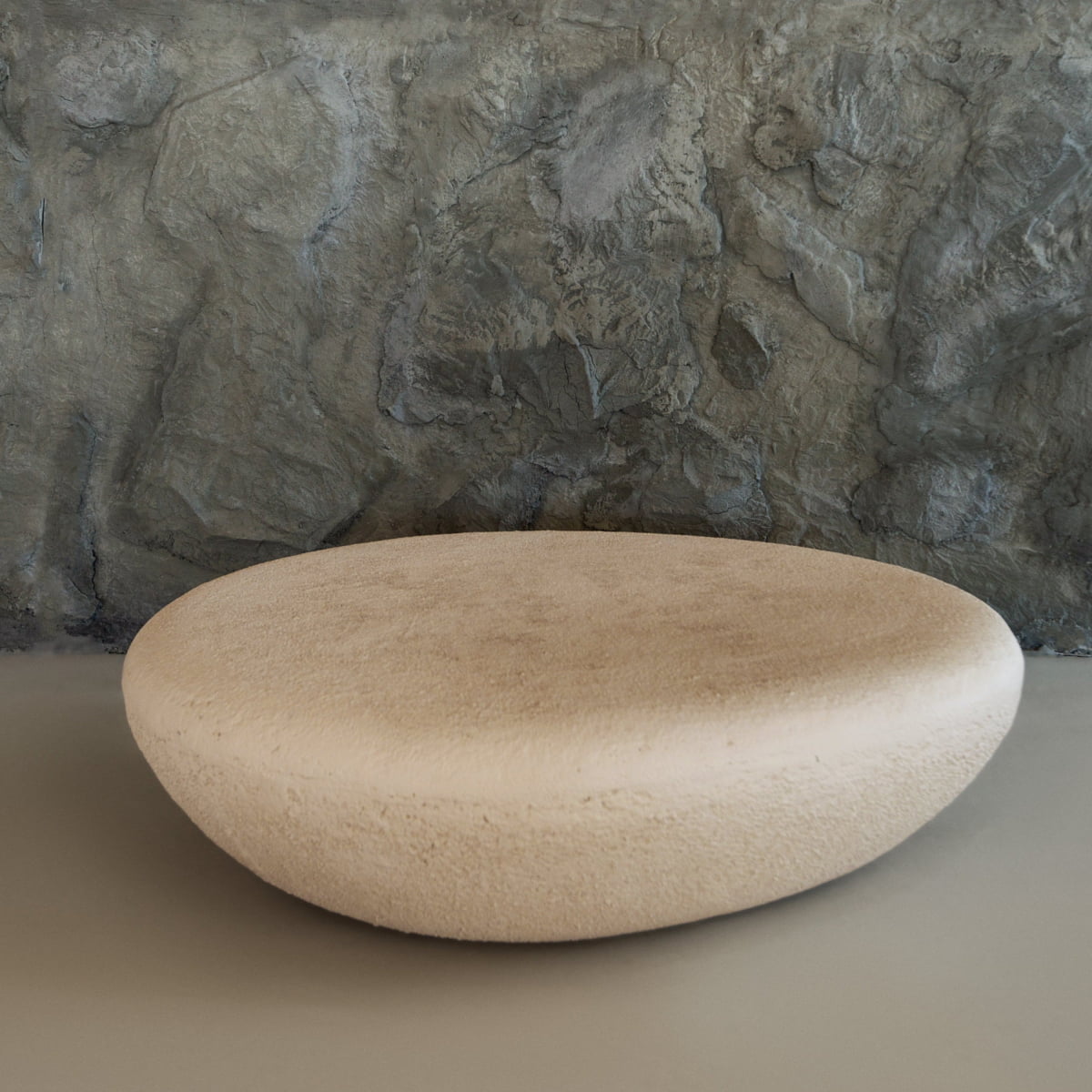 The Pluto Coffee Table features a round, beige, stone-like design that stands out against a textured gray wall, offering modern style and an organic shape—an ideal chic centerpiece for any space.