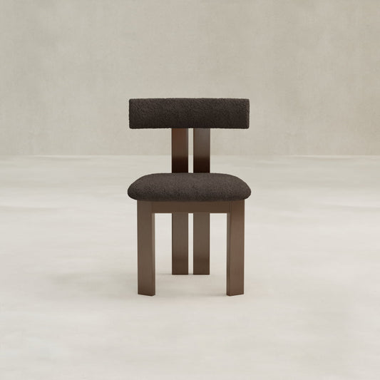 The Radley Dining Chair features a dark, textured bouclé fabric seat and backrest with straight wooden legs, placed on a light floor against a beige wall to provide comfort and modern style.