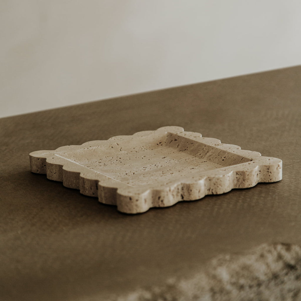 The Remy Tray, featuring a modern square design and scalloped edges in travertine, rests on a textured brown surface with a neutral background.