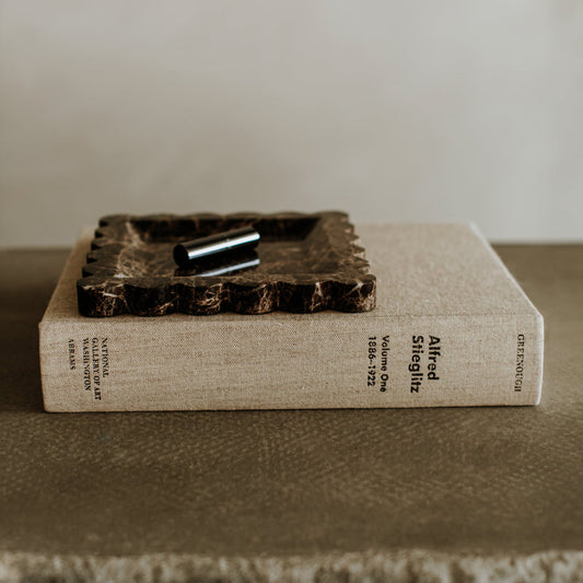 A beige hardcover book labeled Altered Selig 112 and Gestalten sits closed on a taupe surface, topped with the Remy Tray In Marble and a silver cylinder, infusing the scene with modern style.