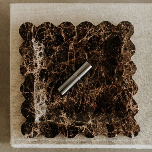 The Remy Tray in Marble, featuring scalloped edges and rich dark brown tones, displays a silver lipstick tube and brings modern elegance to a textured beige linen surface.
