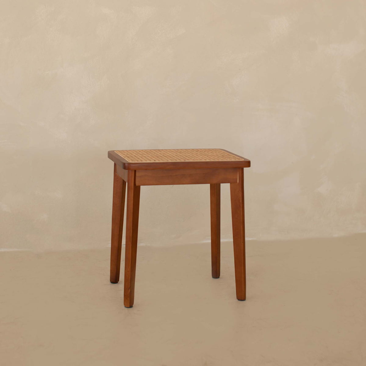 The Rivini Stool features a compact teak wood frame with four legs and a woven rattan seat, shown against a beige textured wall and matching floor.