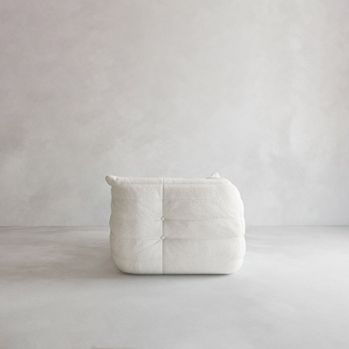 The Rolo Lounge Corner Sofa, featuring a modern, minimalist white upholstered design with boxy tufting and mid-century inspiration, sits alone in a light, neutral-colored room with smooth floors and a plain wall.