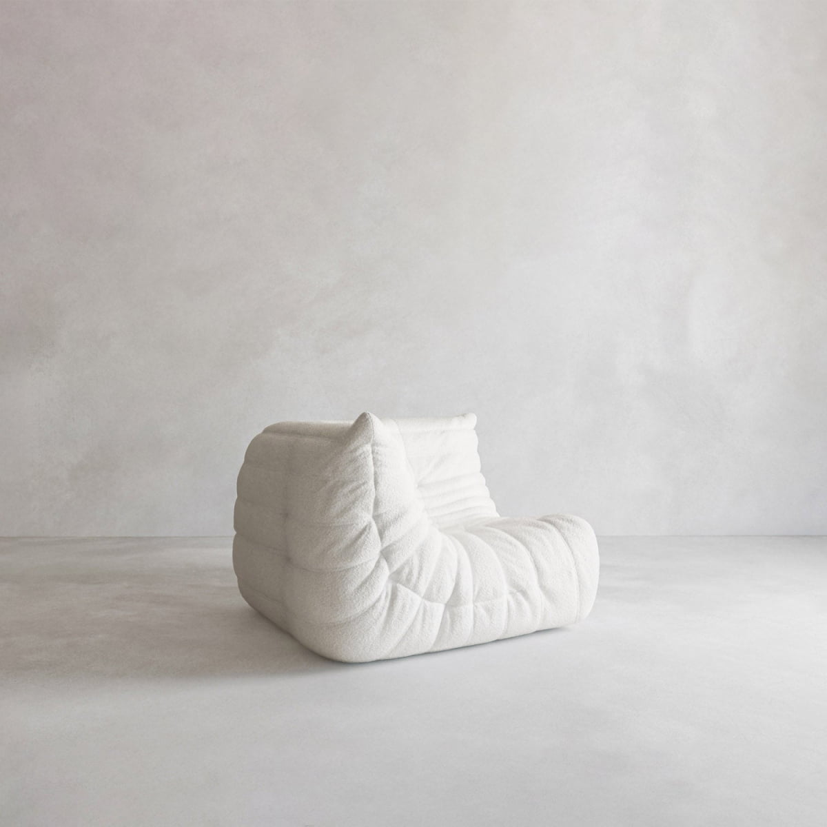 The Rolo Lounge Corner Sofa, a modern, low-profile white piece inspired by the 70s with a quilted tufted design, sits on a plain light floor against a minimalist light gray wall.