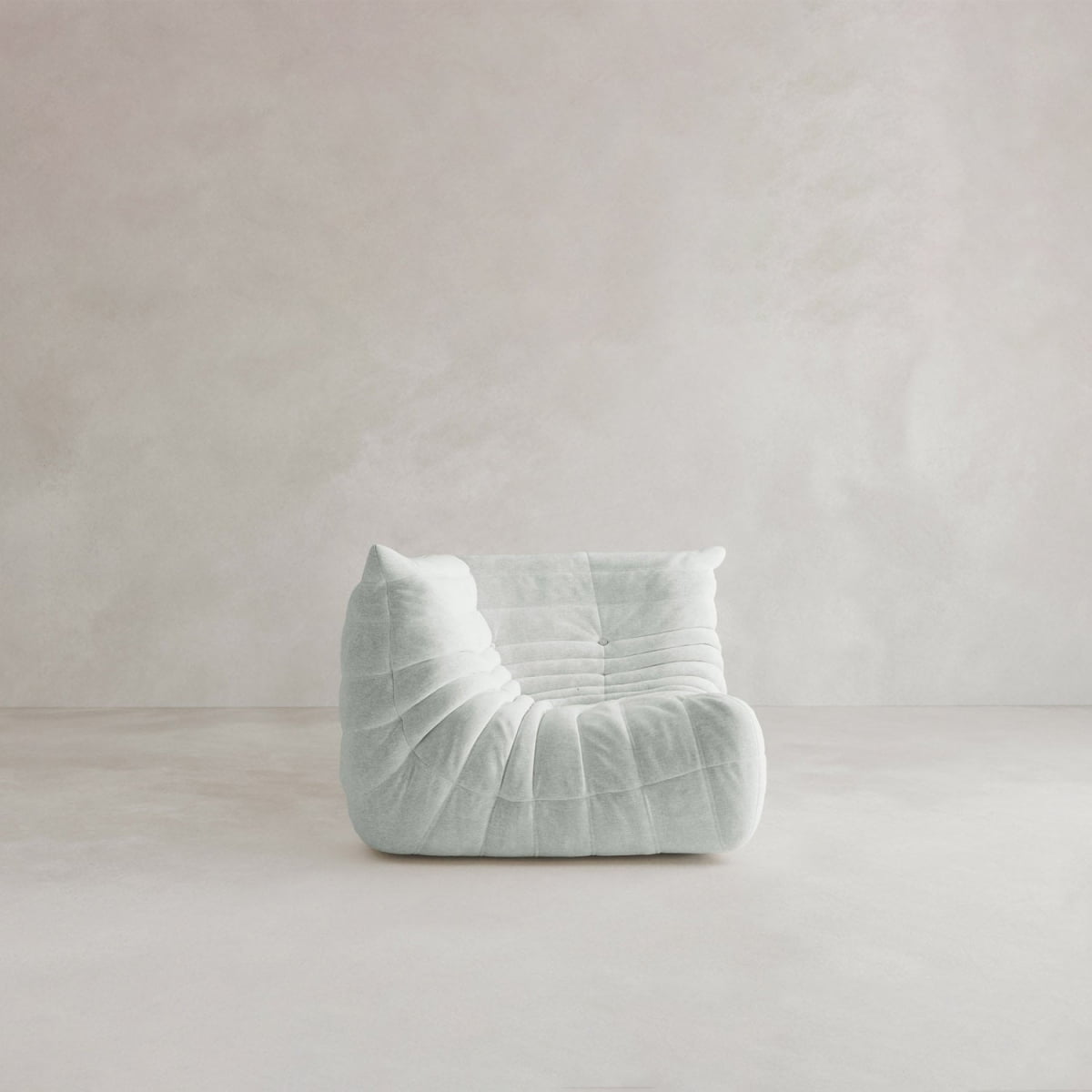 The Rolo Lounge Corner Sofa, featuring soft light grey upholstery and a quilted, ruched design, sits alone in a minimal neutral-toned room with a smooth floor and lightly textured wall.