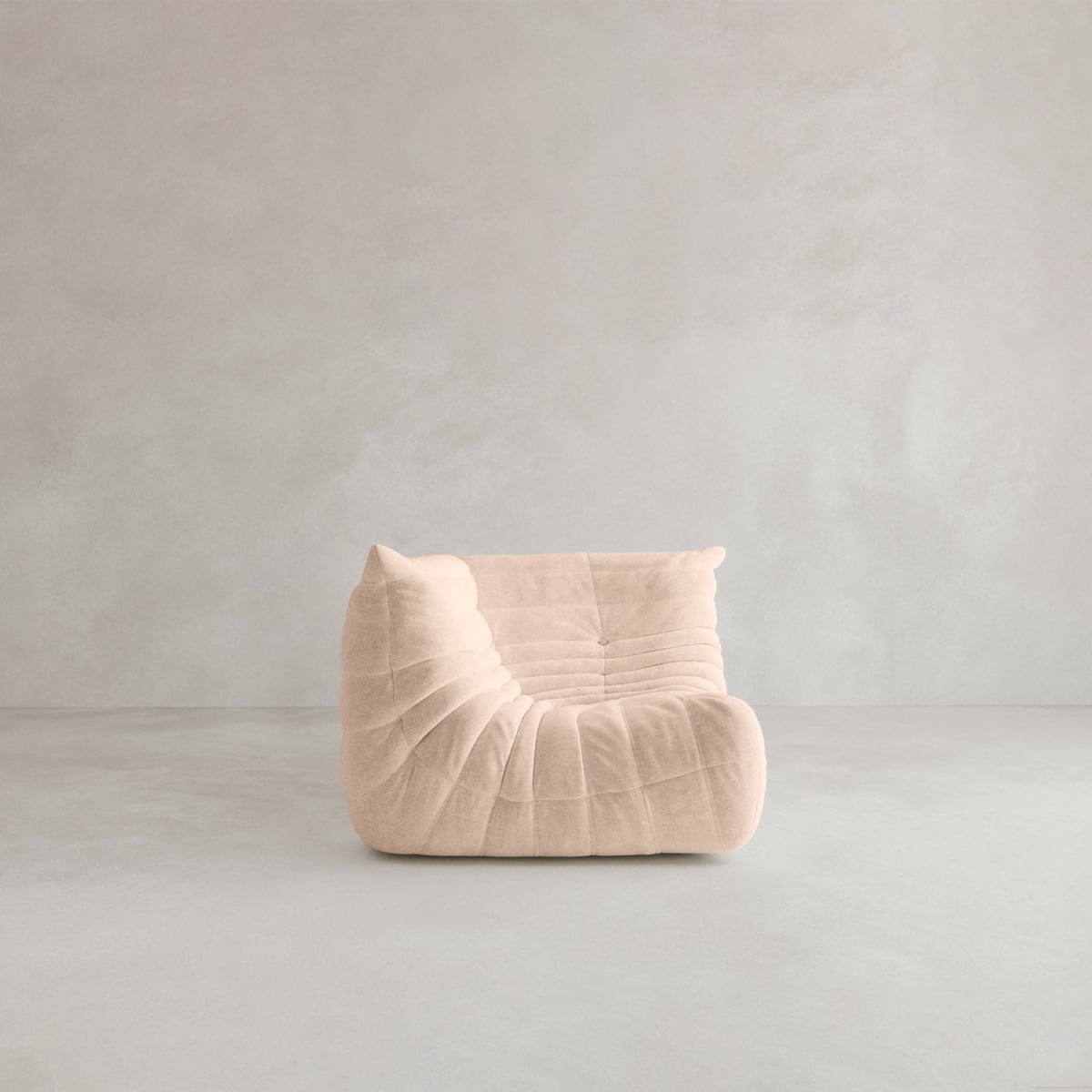 The Rolo Lounge Corner Sofa, a 70s-inspired armless piece in light beige with quilted, ruched upholstery, sits on a plain light gray floor against a matching gray wall.