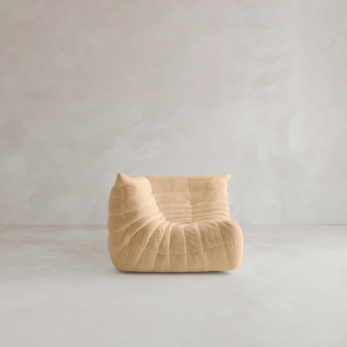 The Rolo Lounge Corner Sofa, a beige armless piece with soft tufted upholstery and subtle mid-century style, sits on a light grey floor against a plain, light-colored wall.