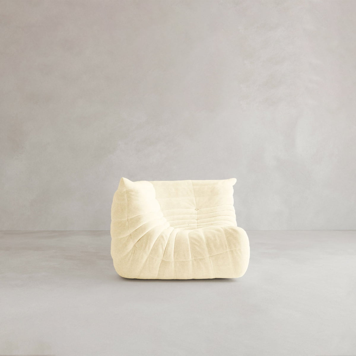 The Rolo Lounge Corner Sofa features modern, cream-colored, armless design with plush, quilted upholstery, capturing a 70s-inspired vibe as it sits centered on a light gray floor against a plain, light gray wall.
