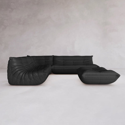 The Rolo Lounge Four Piece Sofa, a modular black sectional with plush tufted cushions, sits in an empty room with light walls and a pale floor, highlighting its sleek mid-century-inspired design.