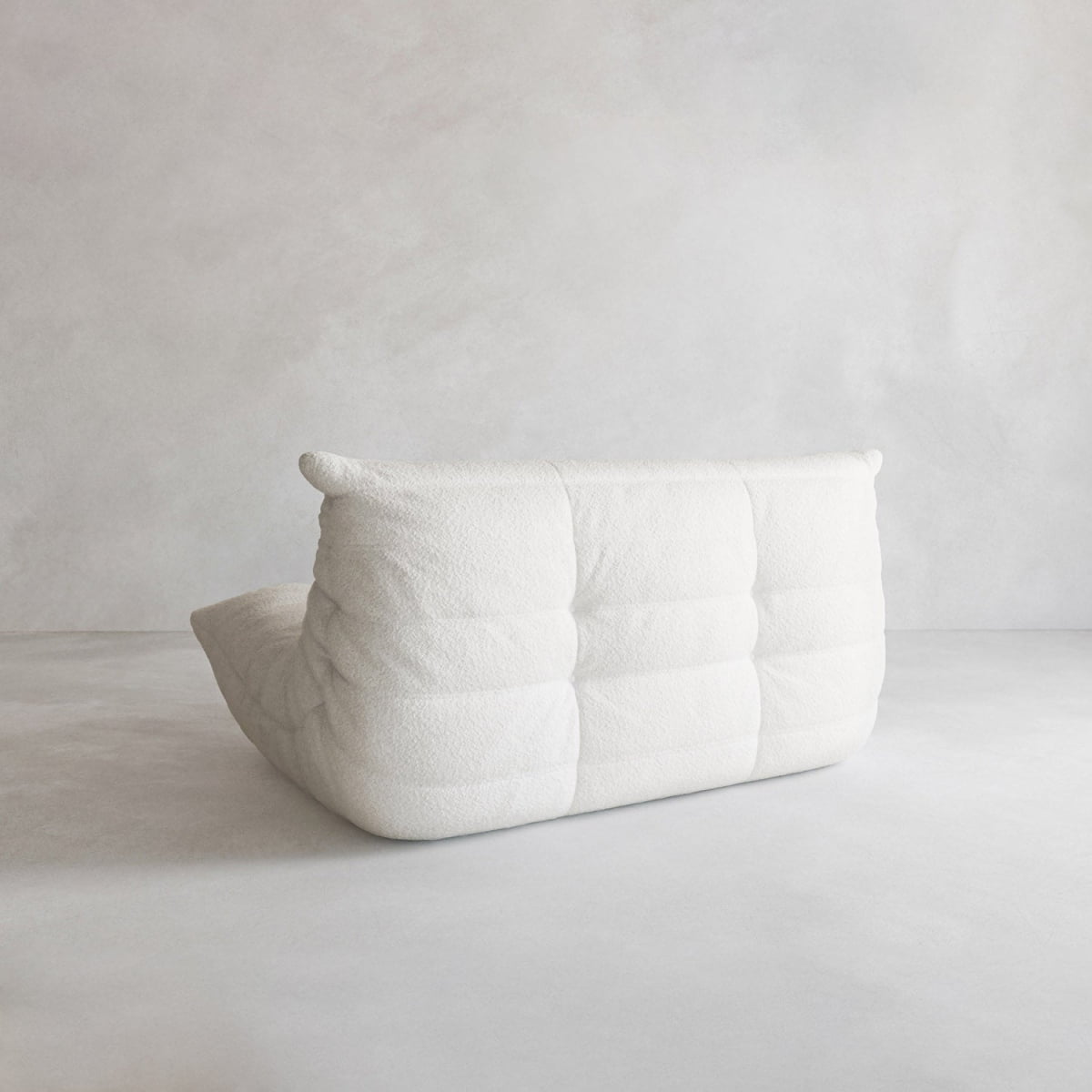 A modern, low-profile, armless Rolo Lounge Loveseat in white with a quilted, textured surface sits on a light floor against a plain light-grey wall, featuring 70s-inspired style and a relaxed, casual look.
