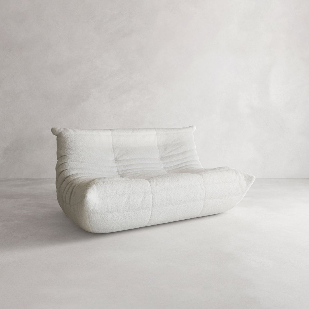 The Rolo Lounge Loveseat, a modern low white upholstered piece with quilted texture and modular design, sits against a plain light gray wall and floor, adding flexible style to the minimalist space.