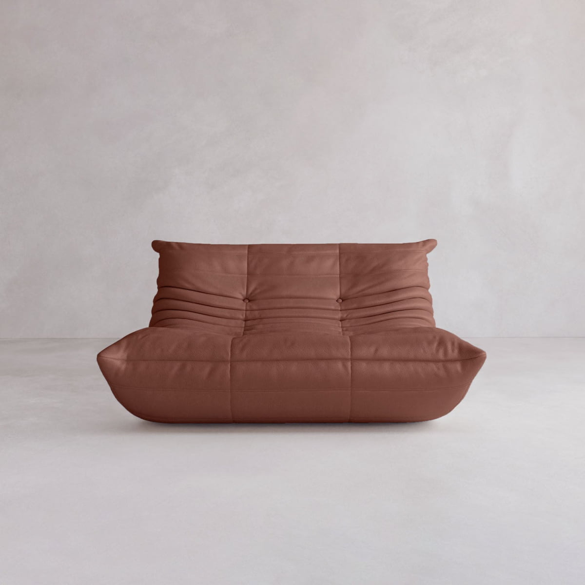 The Rolo Lounge Loveseat, a modern brown leather armless sofa with a low, deep seat and tufted, ruched details inspired by mid-century design, sits against a plain light grey wall and floor.