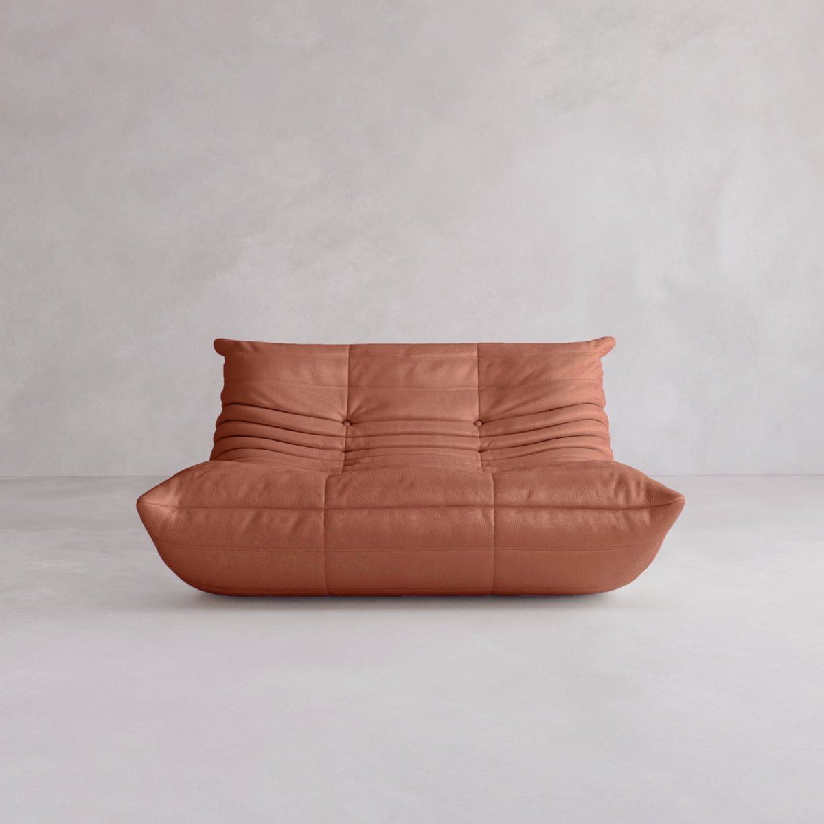 The Rolo Lounge Loveseat features soft, tufted rust-colored upholstery and a modern armless design that evokes mid-century style, sitting low-profile on a light gray minimalist floor against a plain pale wall.