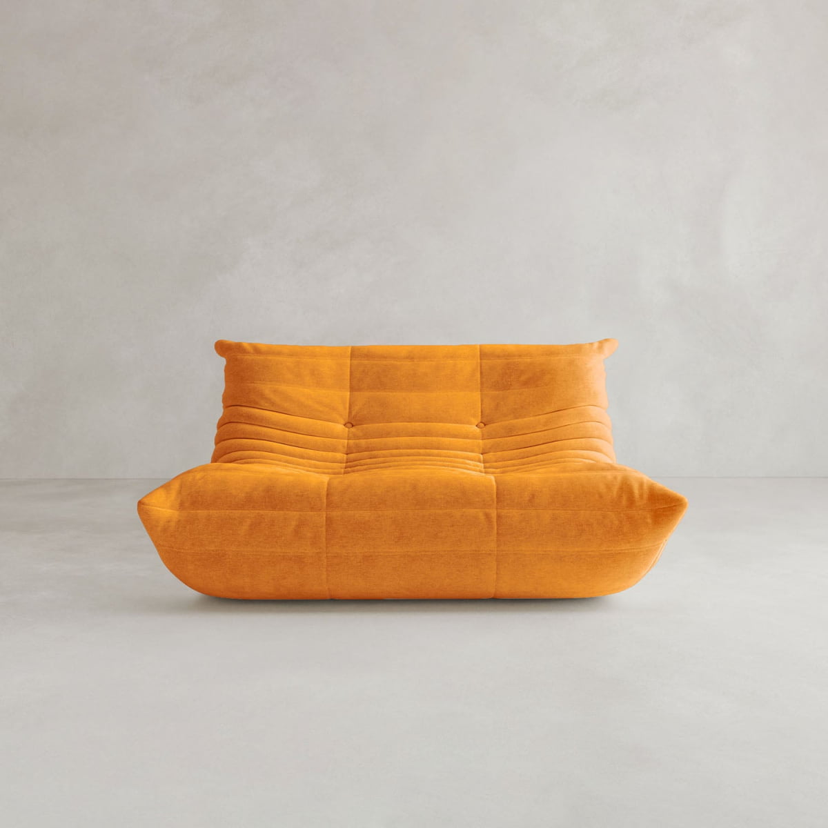 The Rolo Lounge Loveseat, a modern armless orange seat with a low backrest and tufted quilted design inspired by mid-century style, sits on a light grey floor against a plain pale wall.