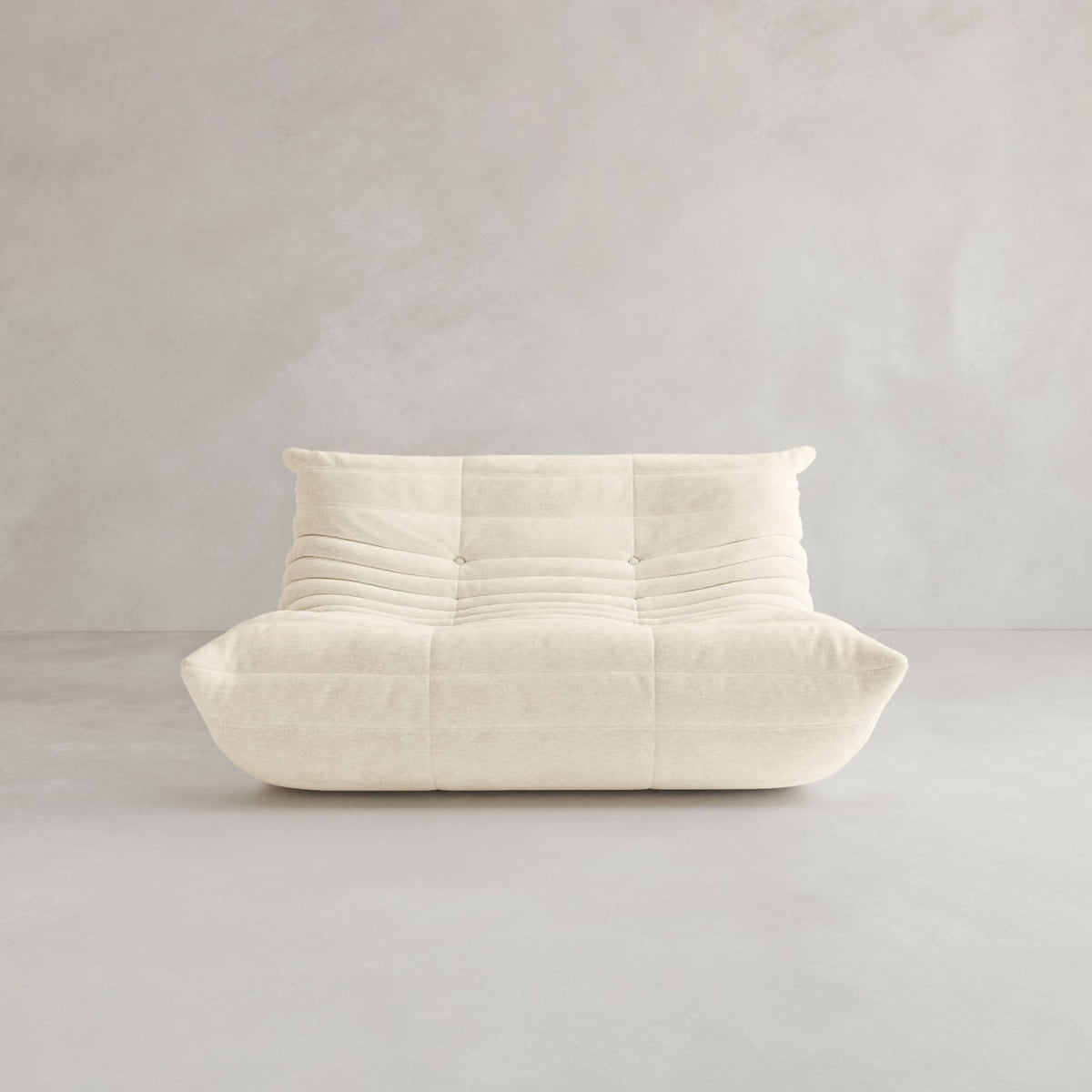The Rolo Lounge Loveseat, a modern armless design with off-white tufted cushions and horizontal stitching, sits against a plain light gray wall and floor, reflecting 70s-inspired style.