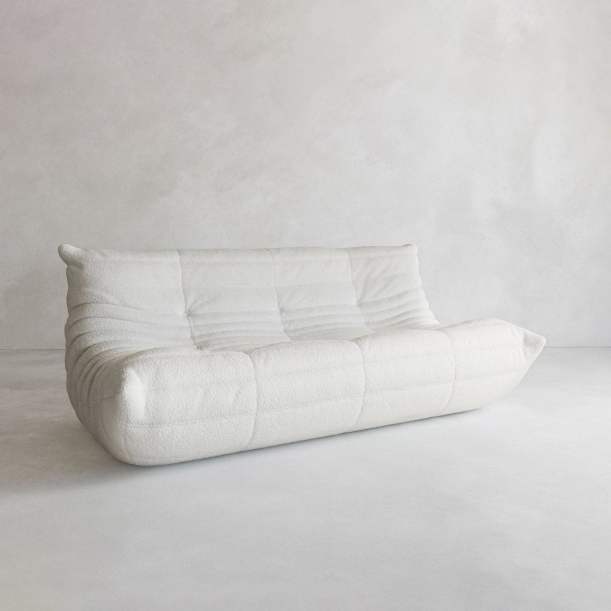 The Rolo Lounge Three Seater is a modern, minimalist white upholstered sofa with a low backrest and soft, rounded edges inspired by mid-century design, shown against a plain light gray wall and floor.