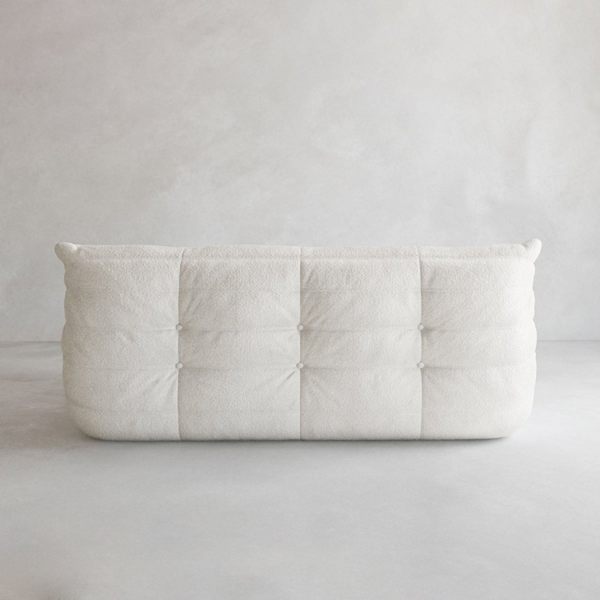 The Rolo Lounge Three Seater, a plush off-white tufted sofa with soft textured fabric, sits against a light gray wall and floor in a minimalist setting, evoking timeless mid-century design.